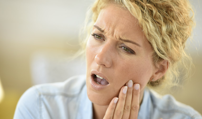 Blonde woman in a blue shirt cringes in pain as she touches her cheek due to extreme tooth pain