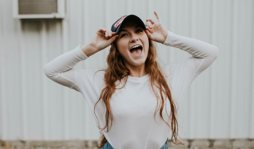 Young woman with long brown hair smiles enthusiastically while wearing a ball cap and white long-sleeved shirt