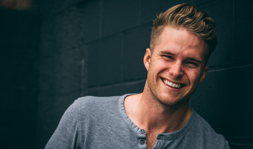 Closeup of a blonde young man smiling against a dark gray wall while wearing a heather gray shirt