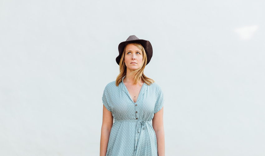 Blonde woman wearing a blue dress and dark hat stands against a gray-blue sky wondering about her teeth color