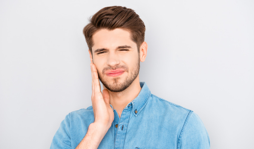 brunette man wearing a denim shirt cringes in pain and touches his cheek due to a toothache