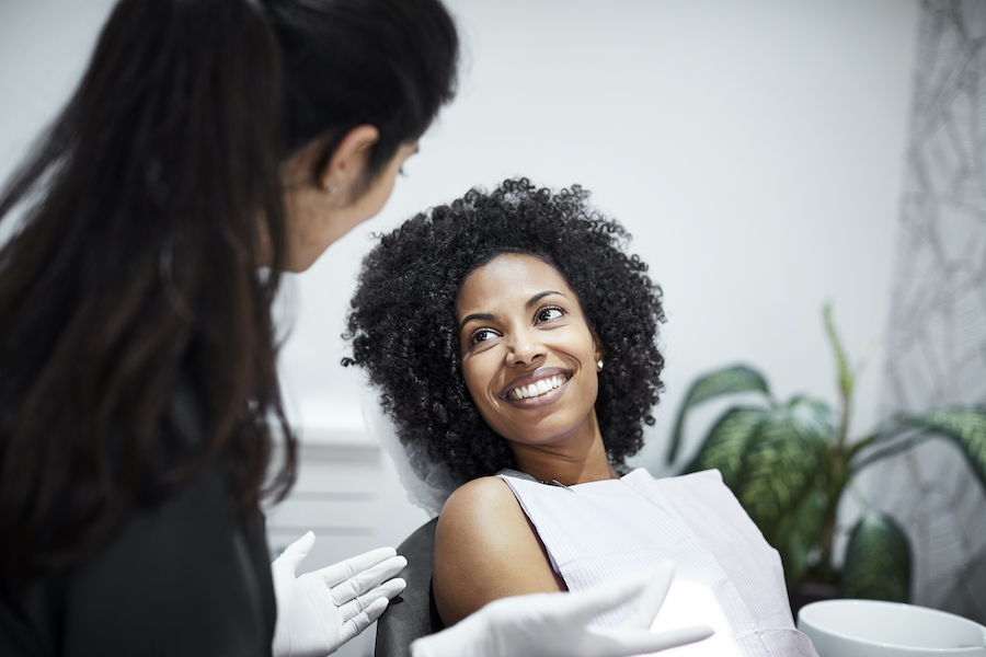 Black woman with curly hair smiles with a white bib covering in a dental chair talking to her dentist