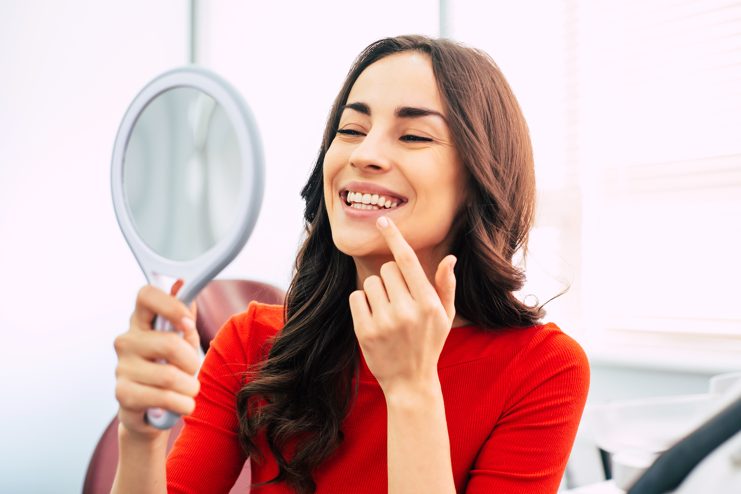 Brunette woman in a red blouse smiles at her teeth in a handheld mirror after professional teeth whitening