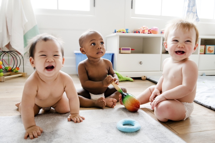 3 babies smile as they sit on the floor together in a playroom