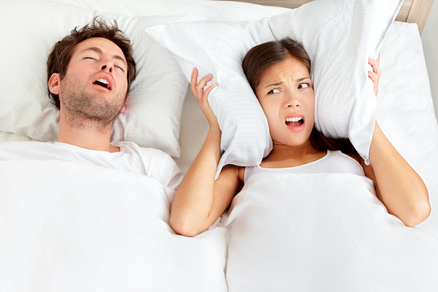 Brunette woman sleeps in bed with his mouth open due to sleep apnea next to his wife who has an annoyed look